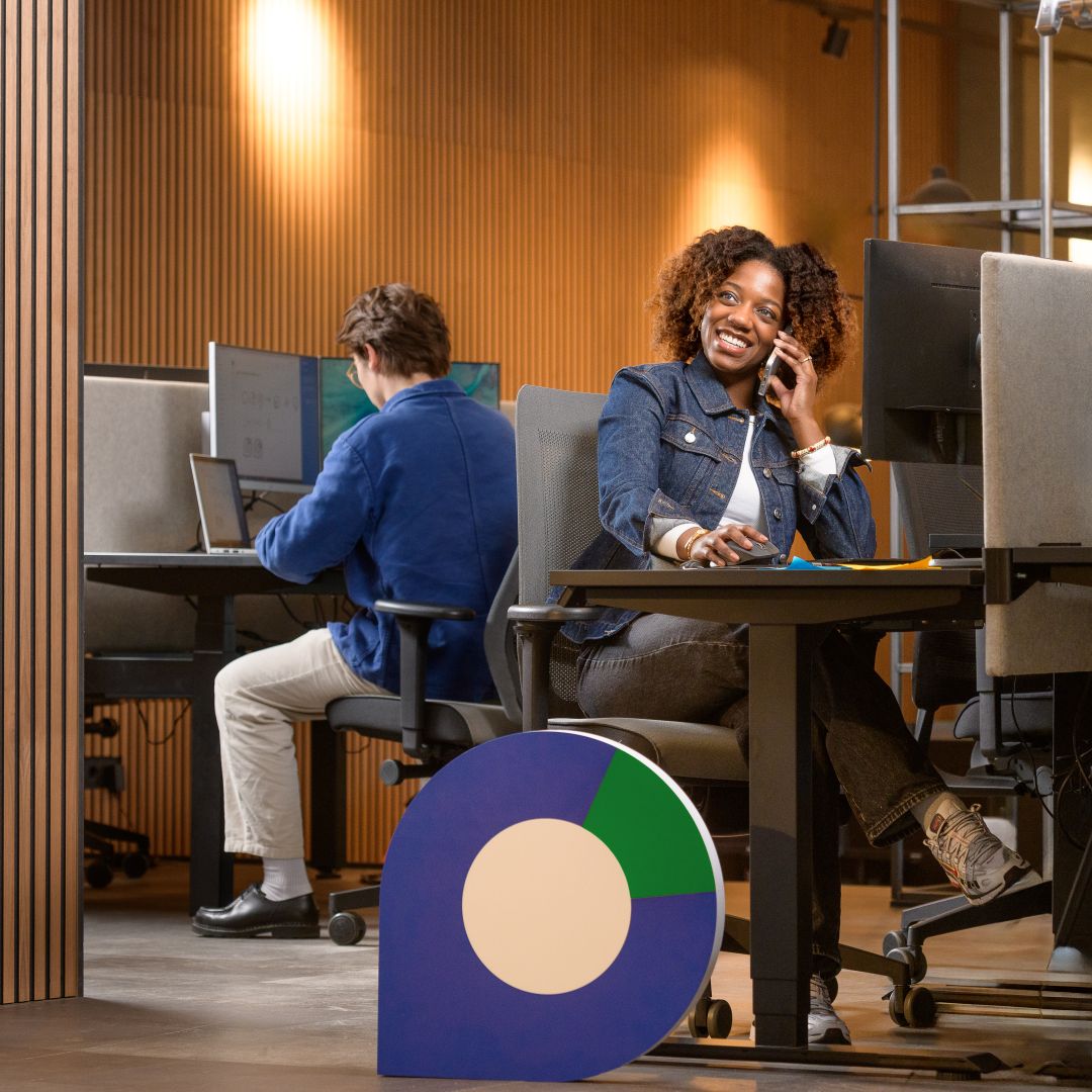 Vrouwelijke hr-adviseur aan het telefoneren achter haar bureau, met daarop een computerscherm. Zij lacht vriendelijk. Naast haar bureau staat een Werkcentrum pointer, regio groen. Achter haar zit een collega achter een laptop en computer te werken.