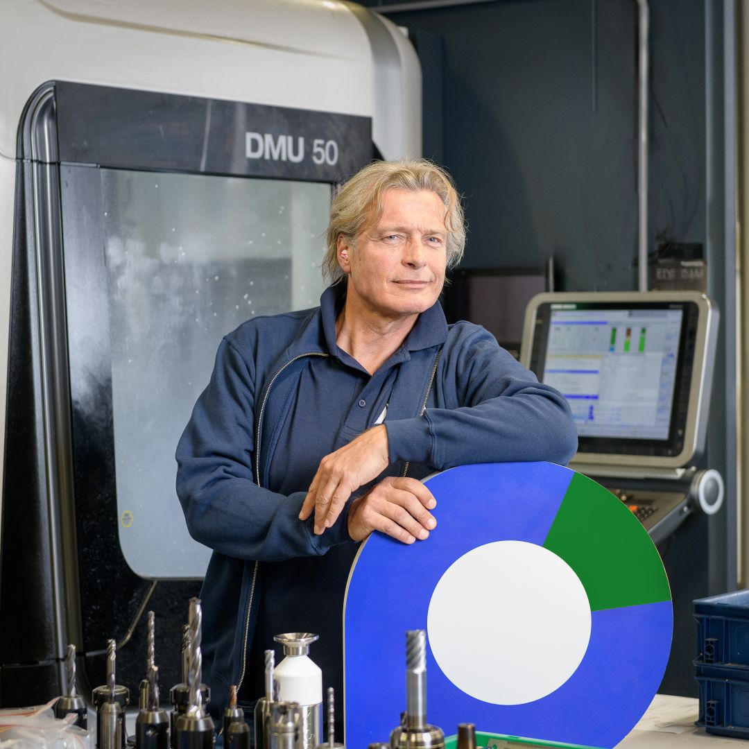 Een mannelijke werkgever in een technische werkplaats staat bij een werkbank en kijkt recht in de camera. Er staan beeldschermen achter hem. De werkgever heeft de pointer in zijn hand en leunt erop met zijn andere arm.