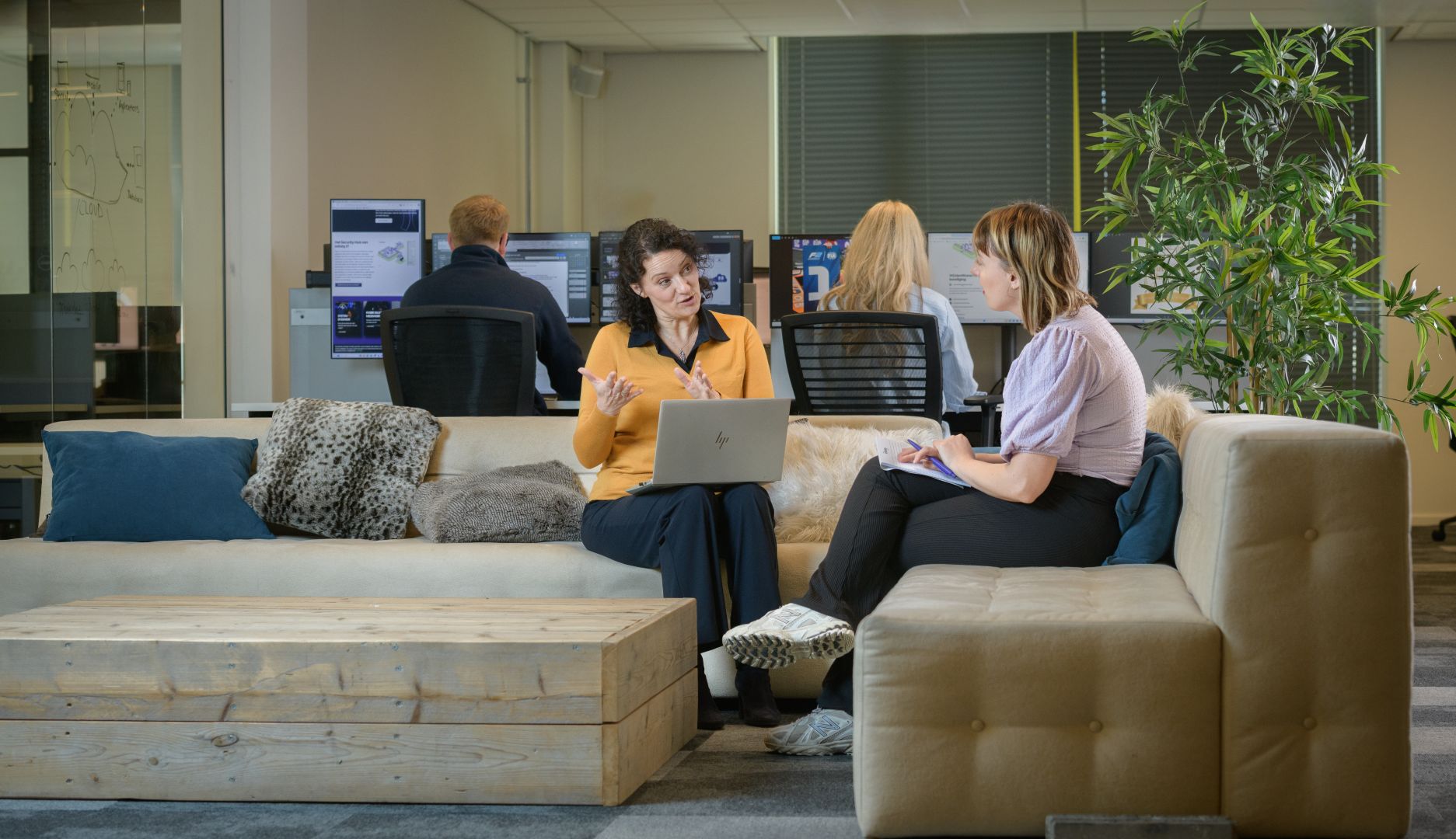 Een vrouwelijke ICT manager zit op een bank te praten met een vrouwelijke collega of klant. Zij zitten op een grote bank en er zitten achter hen ook mensen op computers te werken.