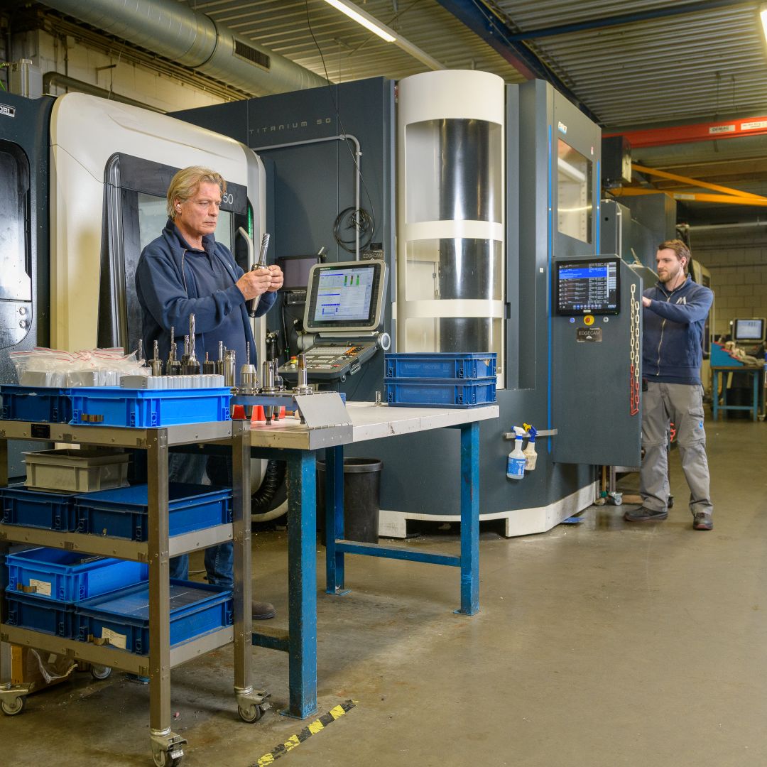 Een mannelijke werkgever in een technische werkplaats staat bij een werkbank. Er staan allerlei bakken met gereedschap om hem heen en er staat een beeldscherm naast hem op de werkbank. Op de achtergrond zie je een jongere werknemer werken aan een soort cabine.